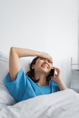 cheerful asian woman with closed eyes touching forehead while talking on smartphone in clinic.