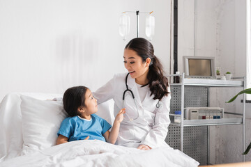 happy asian doctor with stethoscope talking to girl on bed in hospital ward.