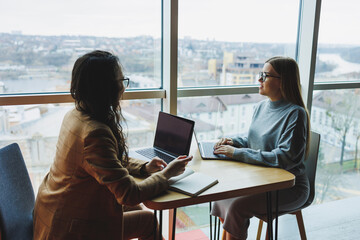 Two young female colleagues sitting with laptops near large windows, modern workspace. Female office workers