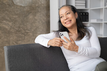 Happy Asain aged woman using her smartphone while sittng on the sofa in the living room.