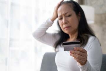 Unhappy Asian senior mature woman holding her credit card, feeling headache and stressed, having financial problems.