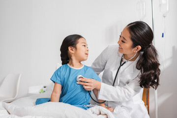 positive doctor in white coat examining asian girl with stethoscope in hospital ward.