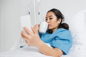 young asian woman drinking tea and looking at blurred cellphone in hospital ward.
