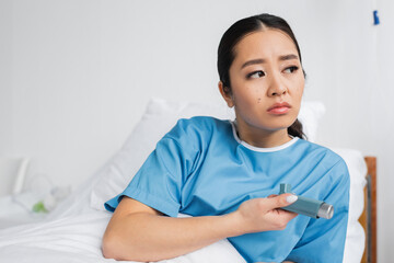 upset and thoughtful asian woman holding inhaler and looking away in hospital.