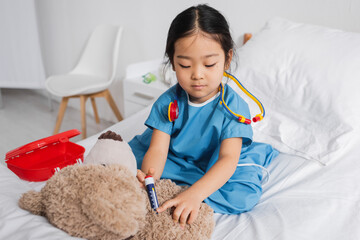 asian child in hospital gown doing injection to teddy bear with toy syringe while playing in clinic.