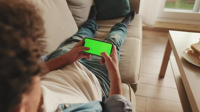 Young man lying on sofa playing on mobile phone with green mock-up screen chroma key