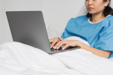 Fototapeta premium partial view of asian woman typing on laptop on bed in hospital ward.