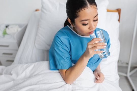 Brunette Asian Woman Sitting On Bed In Hospital Ward And Drinking Fresh Water.