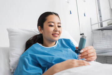 happy asian woman in hospital gown lying on bed in clinic and looking at inhaler.