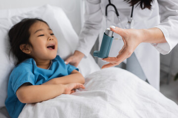 blurred pediatrician holding inhaler near asian child opening mouth on hospital bed.