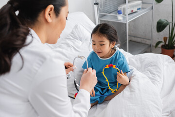 blurred doctor showing stethoscope to positive asian girl sitting with toy on hospital bed.