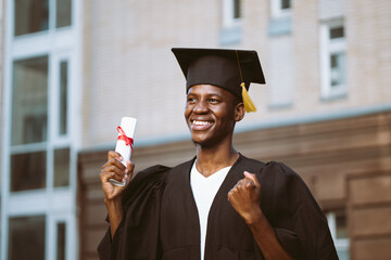 Portrait of smiling black guy graduate from university standing outdoors near building and holding...