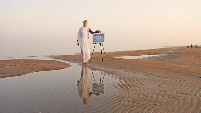 Motion to woman with scarf standing near easel on beach