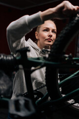 Vertical close-up of focused blonde cycling mechanic female repairing and fixing mountain bicycle standing on bike rack in repair workshop with dark interior. Concept of professional bike maintenance.
