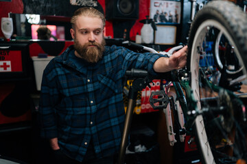 Portrait of pensive bearded cycling mechanic male standing by bicycle in repair bike workshop with dark interior, thoughtful looking away. Concept of professional repair and maintenance of bicycle.