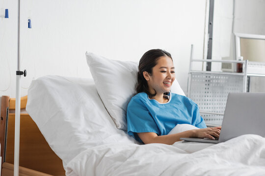 joyful asian woman typing on laptop while staying in hospital bed.