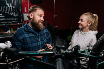 Fototapeta na wymiar Friendly bearded cycling repairman male having conversation with positive smiling blonde female client, talking about problem of bicycle detected during diagnostics, in repair shop with dark interior.