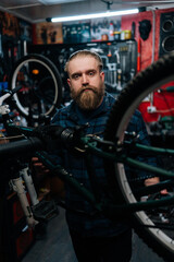 Vertical portrait of brutal cycling repairman standing by bicycle in repair workshop with dark interior, looking at camera. Concept of professional repair and maintenance of bicycle transport.