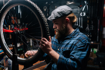 Portrait of serious bearded cycling mechanic male checking bicycle wheel spoke with bike spoke wrench in bike repair shop with dark interior. Concept of professional maintenance of bike transport.