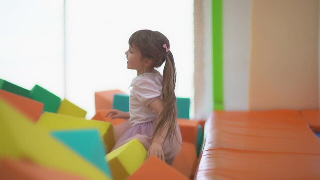 Little happy girl play and have fun in a dry pool with soft multicolored cubes in a children's entertainment center and jump on the paralon blockes in the playroom.