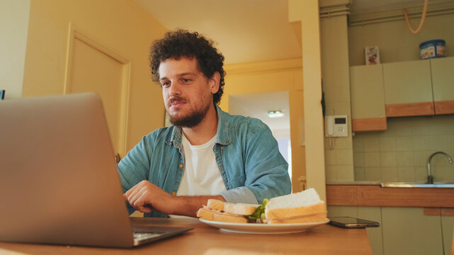 Guy dressed in denim shirt works on laptop while having lunch in cozy kitchen