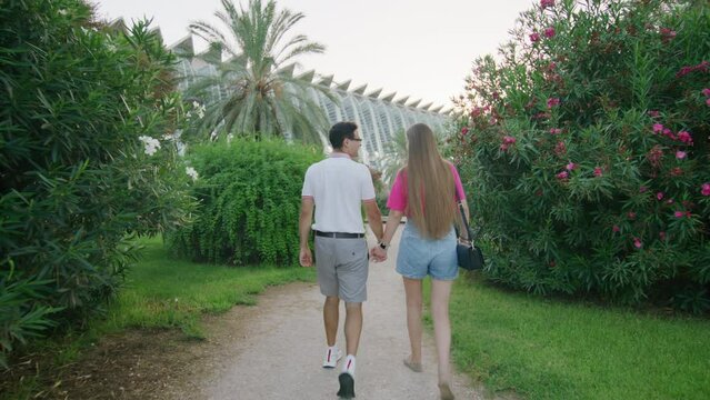 Caucasian boyfriend and girlfriend strolling and feeling in love together. Back view of young couple enjoying summer vacation in charming city park. High quality 4k footage