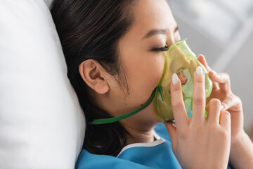 side view of diseased asian woman adjusting oxygen mask in hospital ward.