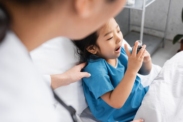 blurred pediatrician touching shoulder of asian child using inhaler in hospital ward.