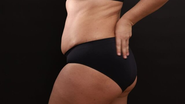 Back view of unrecognizable fat buxom woman wearing black seamless sportswear, adjusting trying on black comfortable sports panties, stretching cloth on black background. Body positive, underwear.