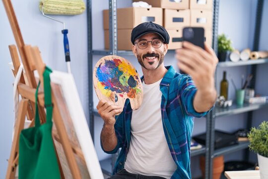 Young hispanic man artist holding palette make selfie by smartphone at art studio