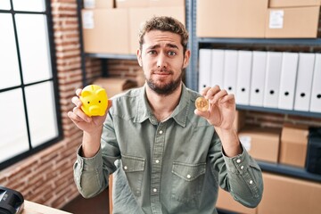 Hispanic man with beard working at small business ecommerce holding piggy bank and bitcoin skeptic and nervous, frowning upset because of problem. negative person.