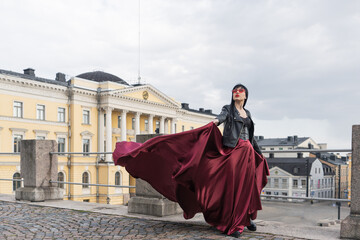 Helsinki. Finland. 25.04.2023 .A sexy woman with a perfect figure stands on Helsinki's Central Square, wearing a red satin skirt and a leather biker jacket. Her skirt billows in the wind