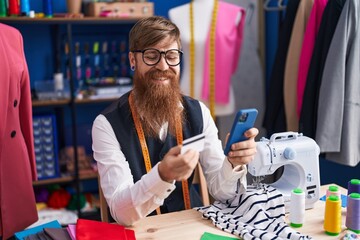 Fototapeta premium Young redhead man tailor using smartphone and credit card at clothing factory