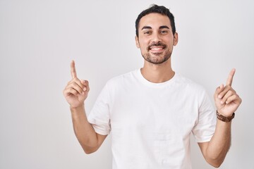Handsome hispanic man standing over white background smiling amazed and surprised and pointing up with fingers and raised arms.
