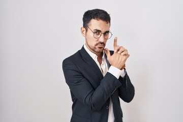 Handsome business hispanic man standing over white background holding symbolic gun with hand gesture, playing killing shooting weapons, angry face