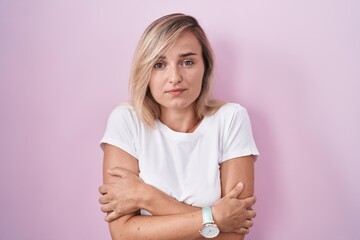 Young blonde woman standing over pink background shaking and freezing for winter cold with sad and...