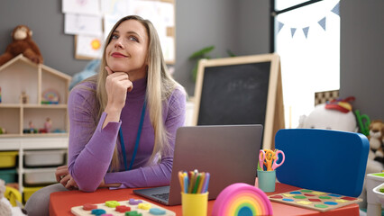 Young blonde woman preschool teacher using laptop sitting on table at kindergarten