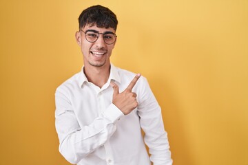 Young hispanic man standing over yellow background cheerful with a smile on face pointing with hand and finger up to the side with happy and natural expression