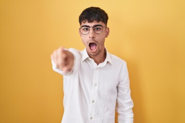 Young hispanic man standing over yellow background pointing with finger surprised ahead, open mouth amazed expression, something on the front