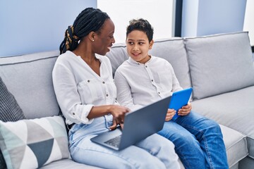 African american mother and son using touchpad and laptop sitting on sofa at home