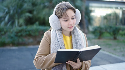 Fototapeta premium Young blonde woman reading book sitting on bench at park