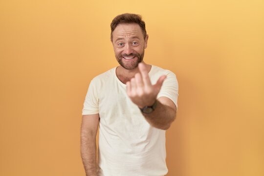 Middle age man with beard standing over yellow background beckoning come here gesture with hand inviting welcoming happy and smiling