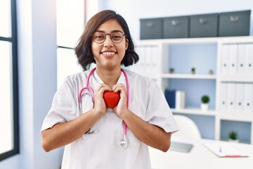 Young latin woman wearing doctor uniform holding heart at clinic