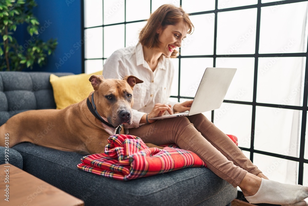 Canvas Prints Young caucasian woman using laptop sitting on sofa with dog at home