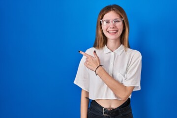 Beautiful woman standing over blue background cheerful with a smile of face pointing with hand and finger up to the side with happy and natural expression on face