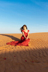 Fototapeta na wymiar Young beautiful woman in long red dress with red rose petals among the desert. Desert rose conception.