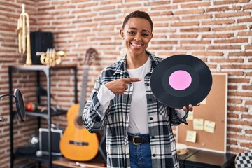 Beautiful african american woman holding vinyl record at music studio smiling happy pointing with...