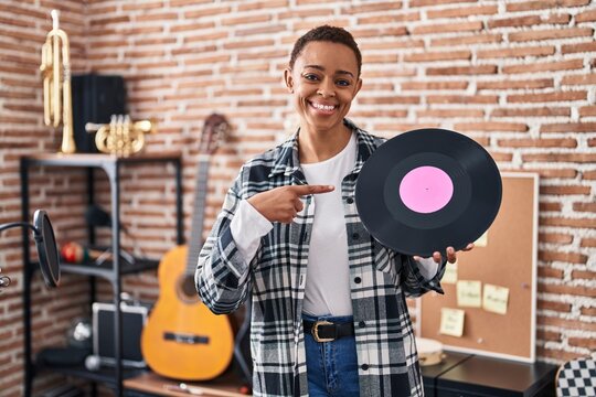 Beautiful african american woman holding vinyl record at music studio smiling happy pointing with hand and finger