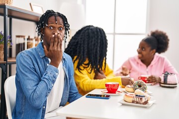 Group of three young black people sitting on a table having coffee covering mouth with hand, shocked and afraid for mistake. surprised expression