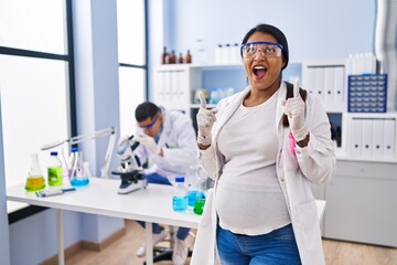 Young hispanic woman expecting a baby working at scientist laboratory amazed and surprised looking up and pointing with fingers and raised arms.